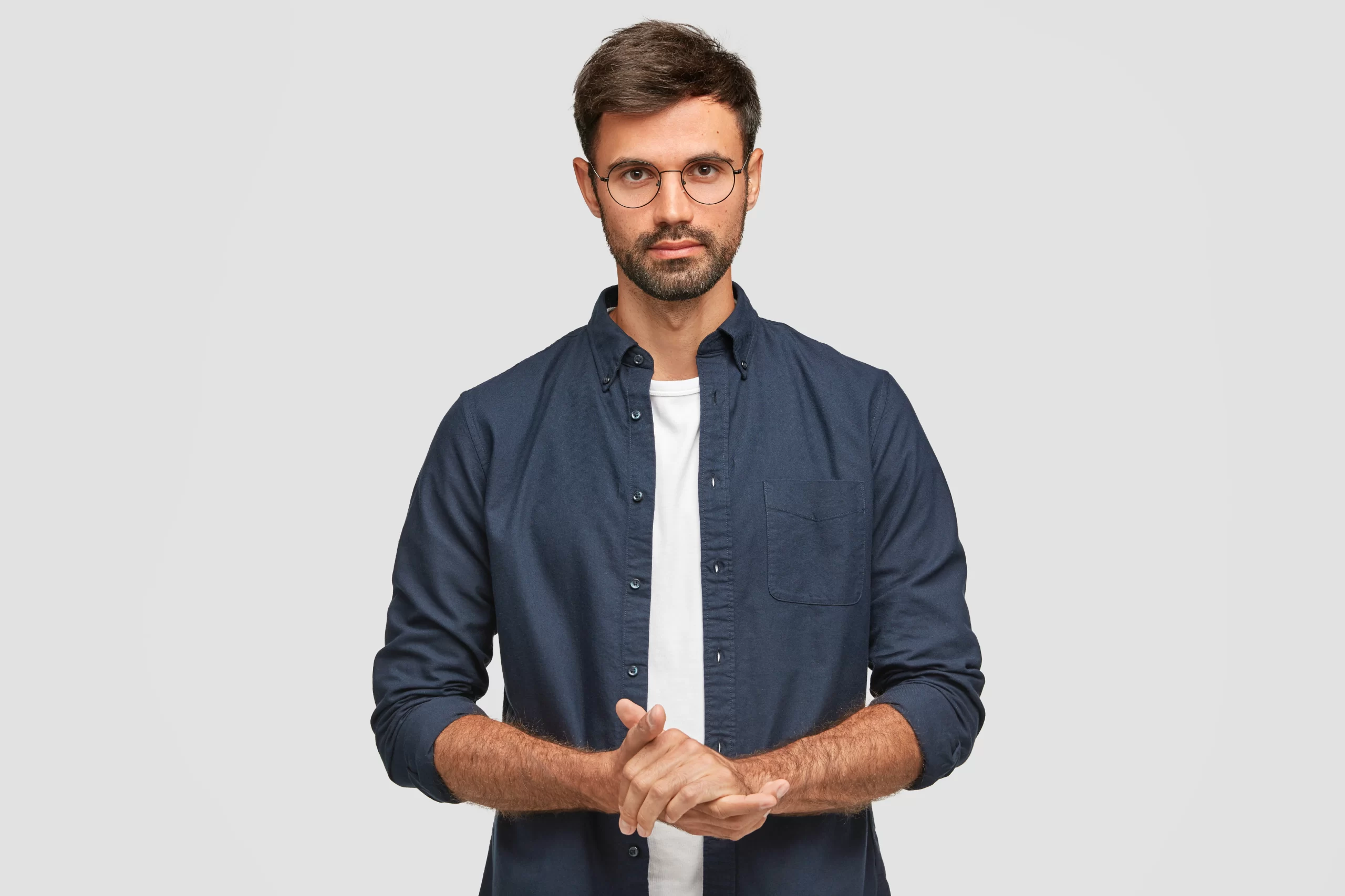 waist-up-portrait-handsome-serious-unshaven-male-keeps-hands-together-dressed-dark-blue-shirt-has-talk-with-interlocutor-stands-against-white-wall-self-confident-man-freelancer
